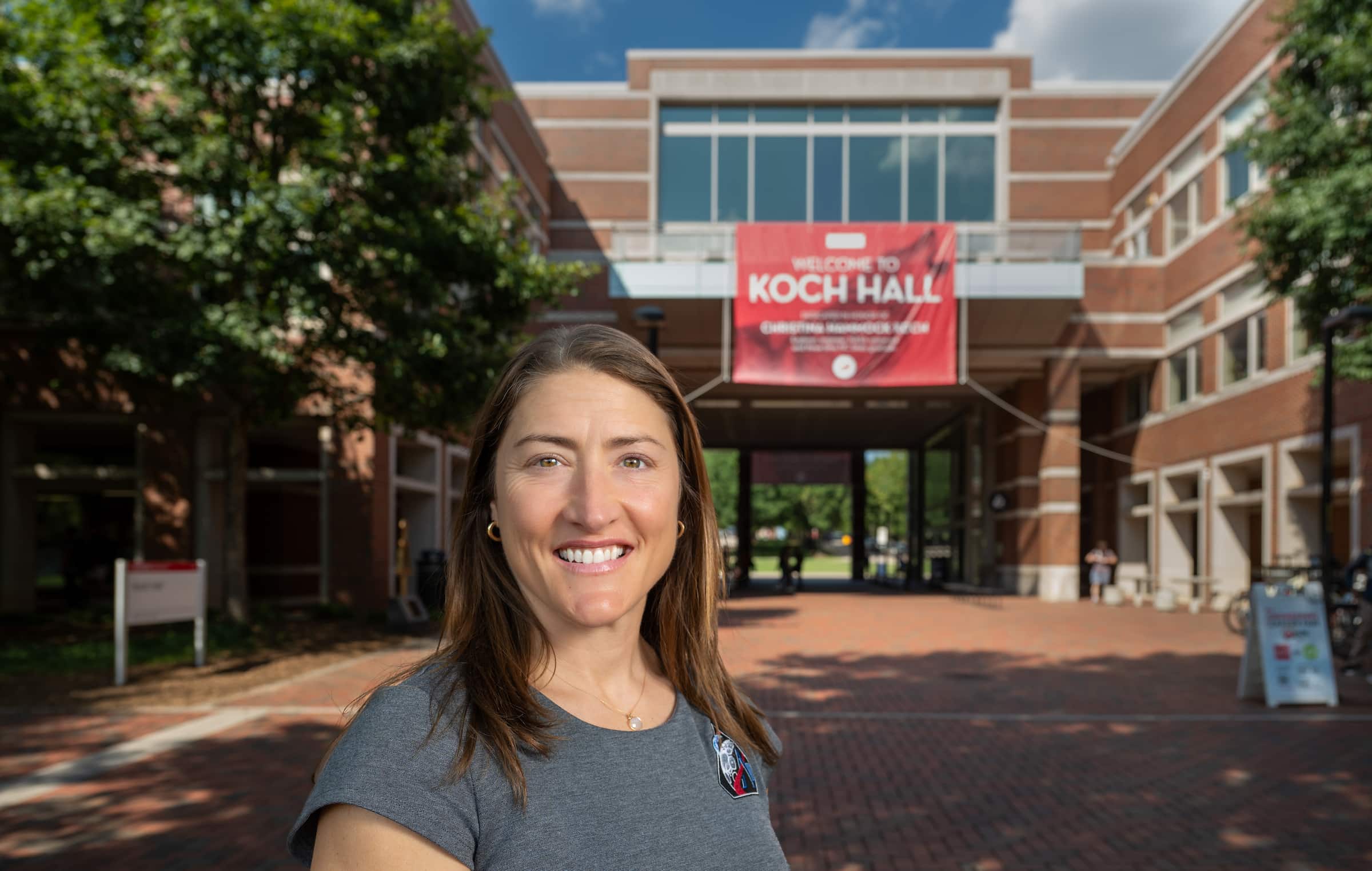 Astronaut Christina Koch stands in front of Koch Hall, with a red banner welcoming visitors to the building on a sunny day.